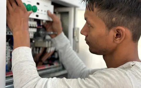 Mężczyzna w jasnej bluzie pracuje przy panelu elektrycznym, montując białe urządzenie z zielonymi przyciskami.