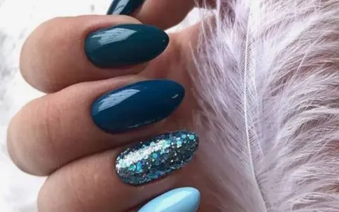 Unghie per l'inverno: unghie blu petrolio e azzurre con glitter scintillanti, accanto a una soffice piuma bianca.