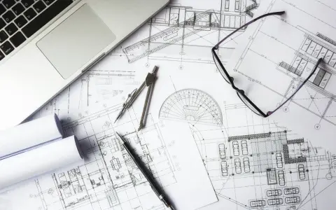 Na stole architekta leżą plany budynku, laptop, okulary i cyrkiel. To miejsce pracy nad projektem i jego kosztorysem.