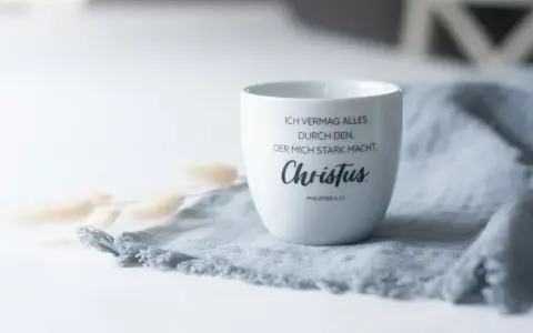 Weiße Tasse mit dem Schriftzug "Ich vermag alles durch den, der mich stark macht, Christus. Philipper 4,13" auf einem grauen Stoff.
