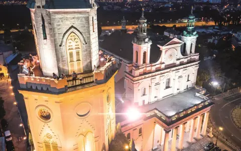 Wieża trynitarska nocą, z ludźmi na balkonie i oświetloną fasadą kościoła.