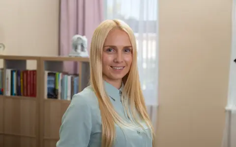 Uśmiechnięta blondynka w jasnej koszuli, za nią regały z książkami.