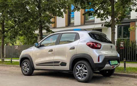 Srebrny Dacia Spring, elektryczny samochód miejski, parkuje pod drzewami. Jego zasięg pozwala na swobodne poruszanie się po mieście.