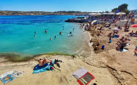 Słoneczna plaża na Malcie w lutym. Ludzie kąpią się w turkusowej wodzie, opalają na piasku i odpoczywają pod parasolami.