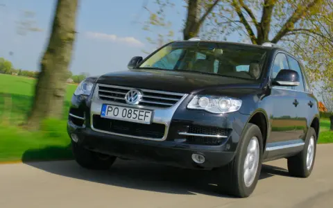 Czarny VW Touareg 3.0 TDI w ruchu, z widocznymi wadami lakieru i drobnymi niedoskonałościami, na tle zielonych pól i drzew.