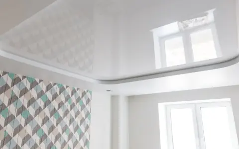 Biały, błyszczący sufit napinany z subtelnymi wzorami geometrycznymi, odbijający światło okna.