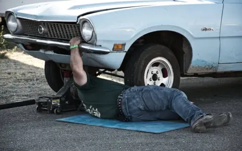 Un uomo lavora sotto un'auto d'epoca, forse per un intervento di **art 46 C.d.S.**, con un cric che la solleva.