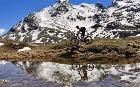 Mountainbiker auf einem epischen Alpencross-Abenteuer, die schneebedeckten Gipfel spiegeln sich in einem Bergsee.
