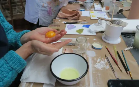 Préparation d'une tempera peinture à l'oeuf : un jaune d'œuf est tenu dans une main au-dessus d'un bol contenant le blanc.