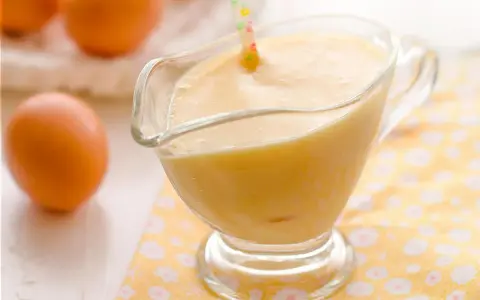 Crema pastelera cremosa en una salsera de cristal, con huevos frescos al fondo.