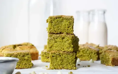 Grüne, saftige matcha brownies, gestapelt und auf einem Tisch arrangiert, mit weißen Schokostückchen und Milchflaschen im Hintergrund.