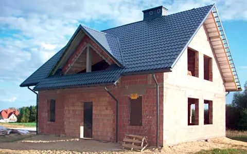 Budowa domu z cegły, widoczny dach betonowy skośny ocieplenie. Widać otwory okienne i drzwiowe, komin.