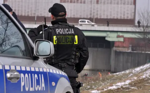 Policjant w kamizelce z napisem "POLICJA", zgodnej z rozporządzeniem w sprawie umundurowania policjantów, stoi obok radiowozu.