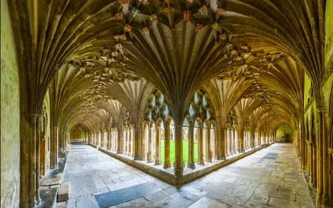 A grand cloister with vaulted ceilings, reminiscent of a monastery floor plan. Sunlight streams through arched windows onto the stone floor.