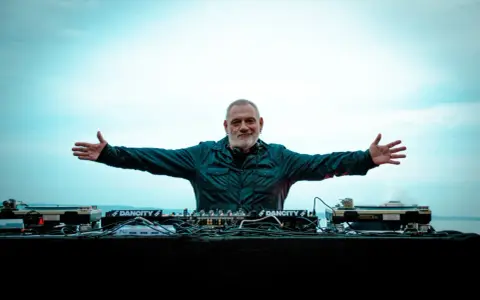 DJ Ralf, un vero patrimonio della musica elettronica, con le braccia aperte davanti alla sua consolle, pronto a far ballare il mondo.