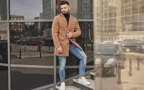 Mężczyzna w czarnym golfie i beżowym płaszczu stoi na schodach.