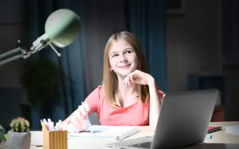 Uśmiechnięta dziewczynka z długopisem w dłoni, zastanawia się nad zadaniem. Obok laptop, dlatego jaka to część mowy?