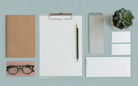 Un exemple de book artistique : un carnet brun, des lunettes, un presse-papiers avec un crayon, un carnet gris, une plante verte, des cartes de visite et une enveloppe.