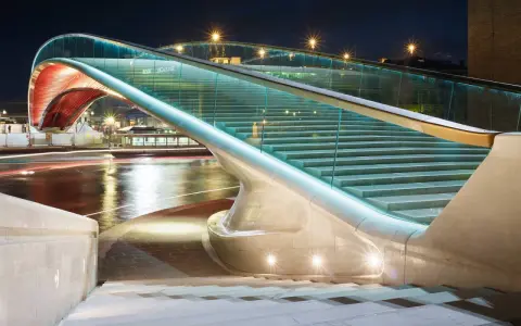 Le pont de la Constitution, illuminé la nuit, avec ses escaliers de verre et ses arches rouges, surplombe un canal aux reflets lumineux.