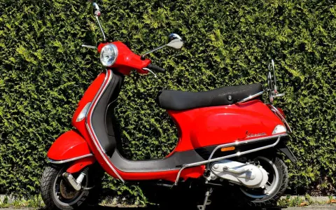 Scooter rojo brillante aparcado frente a un seto verde. ¡Listo para salir con mi permiso AM!
