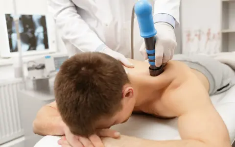 Arzt behandelt HWS-Syndrom mit Stoßwellentherapie. Ein Mann liegt auf dem Bauch, während ein Arzt mit einem blauen Gerät auf seinen Rücken drückt.