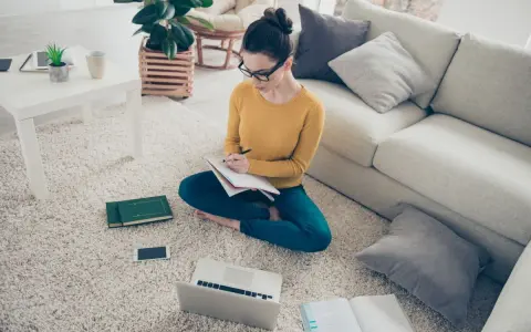 Dziewczyna w okularach uczy się w domu, przygotowując się do egzaminów licealnych. Na dywanie ma laptopa, książki i notatnik.