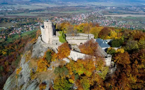 Zamek Chojnik na skalistym wzgórzu, otoczony jesiennymi barwami drzew. W oddali widać miasteczko i pola.