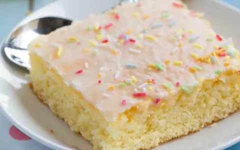 Ein Stück saftiger Fantakuchen vom Blech, mit Zuckerguss und bunten Streuseln bestreut, auf einem Teller mit Löffel.