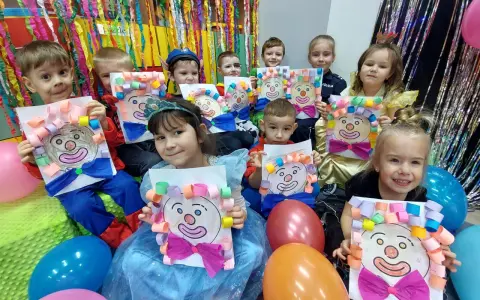 Dzieci w przedszkolu cieszą się z zabaw na balu karnawałowym, prezentując własnoręcznie wykonane prace z wizerunkami klaunów.