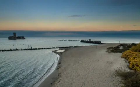Zachód słońca nad morzem w Gdyni. Na wodzie widać betonową fortecę, a na brzegu piaszczystą plażę i babie doły.
