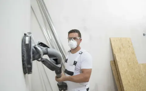 Operaio con maschera e occhiali sta per scartavetrare muro con una levigatrice.