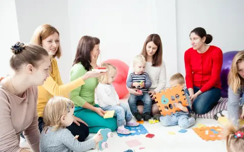 Grupa dzieci i dorosłych uczestniczy w zajęciach, które przypominają terapię pedagogiczną. Dzieci bawią się klockami i zabawkami.