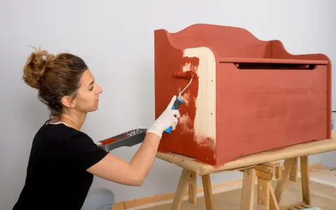 Mujer con guantes blancos pinta con rodillo una caja de madera de color terracota.