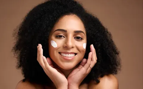 Une femme souriante applique du beurre de karité visage sur ses joues. Ses mains caressent doucement sa peau.