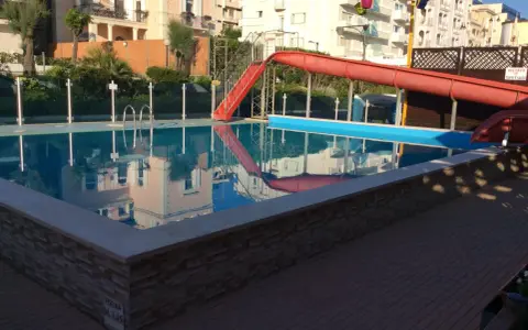 Piscina con scivolo rosso e giochi d'acqua ai Bagni Carmen Cattolica. Riflessi degli edifici nell'acqua azzurra.