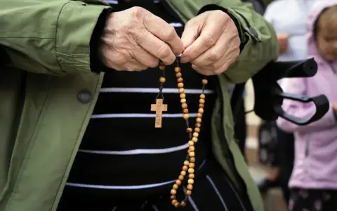 Dłonie starszej osoby trzymające różaniec. W tle widać inne osoby, w tym dziecko. To modlitwa o pracę dla syna.