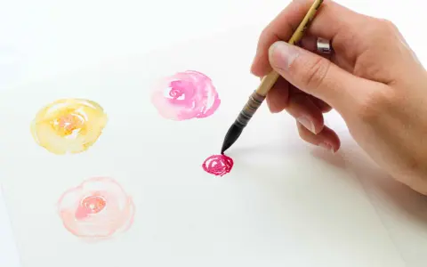 Un dessin de champs de fleurs facile à l'aquarelle. Une main peint des roses délicates en jaune, rose et rouge sur du papier blanc.