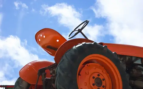 Close-up of an orange tractor's wheel and seat against a blue sky with clouds. This vintage tractor, possibly a different size Kubota tractor, awaits its next task.