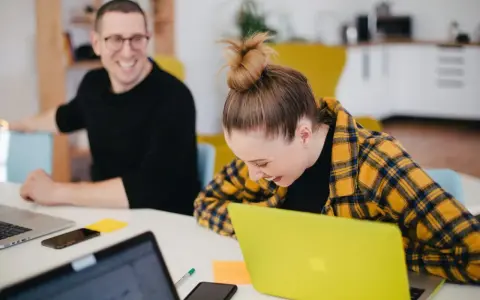 Zwei lachende Kollegen am Schreibtisch mit Laptops. Ein bezahltes Auslandspraktikum kann so Spaß machen!