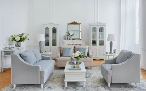 Salotto in stile shabby chic con divani grigi, poltrone, tavolino bianco, specchio dorato e vetrine bianche.