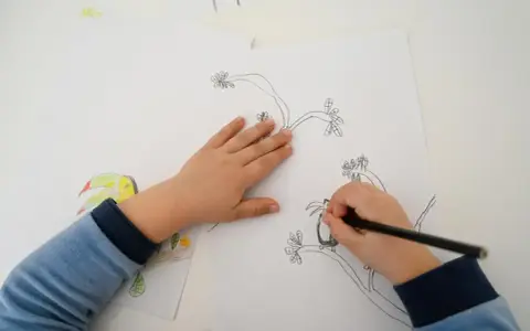 Dziecko rysuje ołówkiem papugę na gałęzi. Obok widać już kolorowy rysunek tukana.
