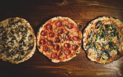 Drei italienische Pizzen: eine mit Pilzen, eine mit Salami und eine mit Gemüse, liegen auf einem Holztisch.