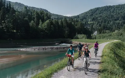 Gruppe Radfahrer auf einem Weg entlang eines Flusses mit bewaldeten Hügeln im Hintergrund.