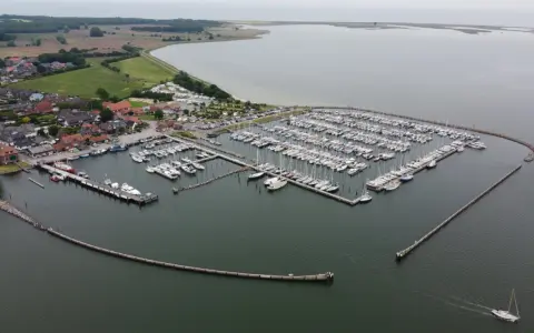 Maasholm Hafen mit vielen Segelbooten und Yachten, die an Stegen liegen. Im Hintergrund ein Dorf und das weite Wasser.