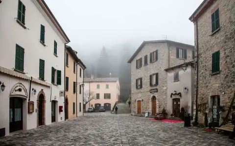 Piazza di San Leo avvolta nella nebbia, con edifici in pietra e intonaco, un bar e una farmacia.