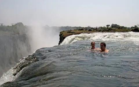 Zwei Personen schwimmen im Devils Pool am Rande der Victoriafälle, mit Gischt und üppiger Vegetation im Hintergrund.