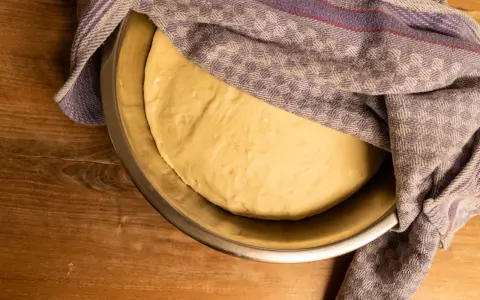Pizzateig ruht in einer Schüssel, bedeckt mit einem gemusterten Tuch. Die Frage "wie lange pizzateig gehen lassen" beschäftigt den Bäcker.