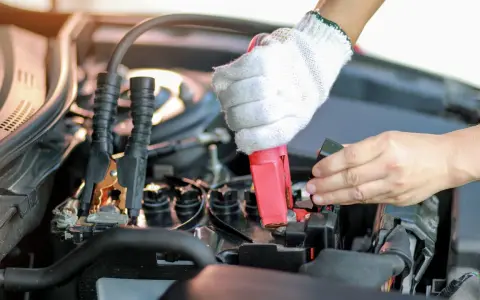 Eine Hand mit Handschuh klemmt das rote Kabel an den Pluspol, während die andere Hand den Minuspol der Autobatterie sichert.