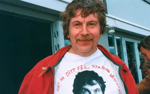 Staffan Westerberg, känd för sina filmer och tv-program, ler i en röd jacka över en t-shirt med hans eget ansikte och texten "Det är ditt fel, Staffan Westerberg".