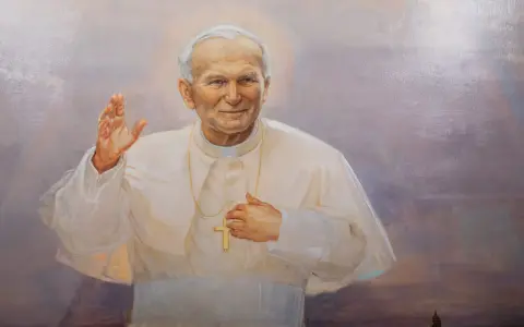 Obraz przedstawia Jana Pawła II w białej szacie, z podniesioną prawą dłonią i krzyżem na piersi. Wierni odmawiają modlitwę do Jana Pawła II o uzdrowienie.